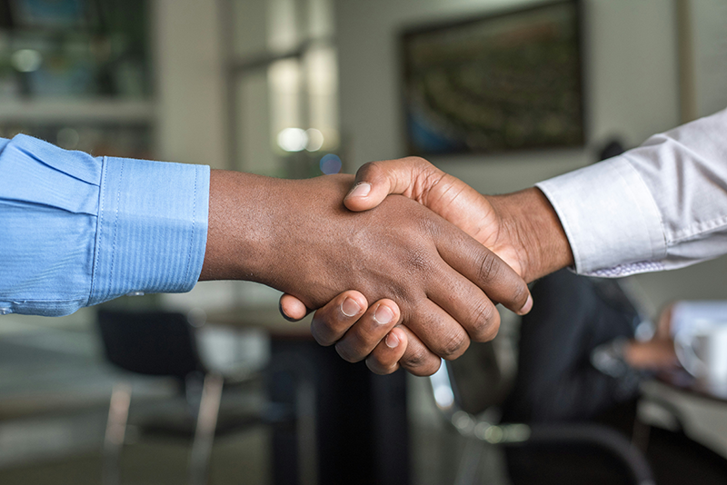 a set of hands shaking. One person is wearing a light blue long sleeved cuffed shirt. The other person is wearing a grey long sleeved cuffed shirt. An office space is behind them showing desks and chairs, blurred.