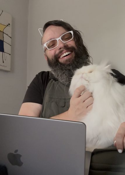 Dylan working from home, sitting on his bed in front of his laptop, holding his very fluffy white cat, Blue.